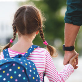 A young girl holding her father's hand as she goes to school. She is wearing a pink shirt and a blue backpack.